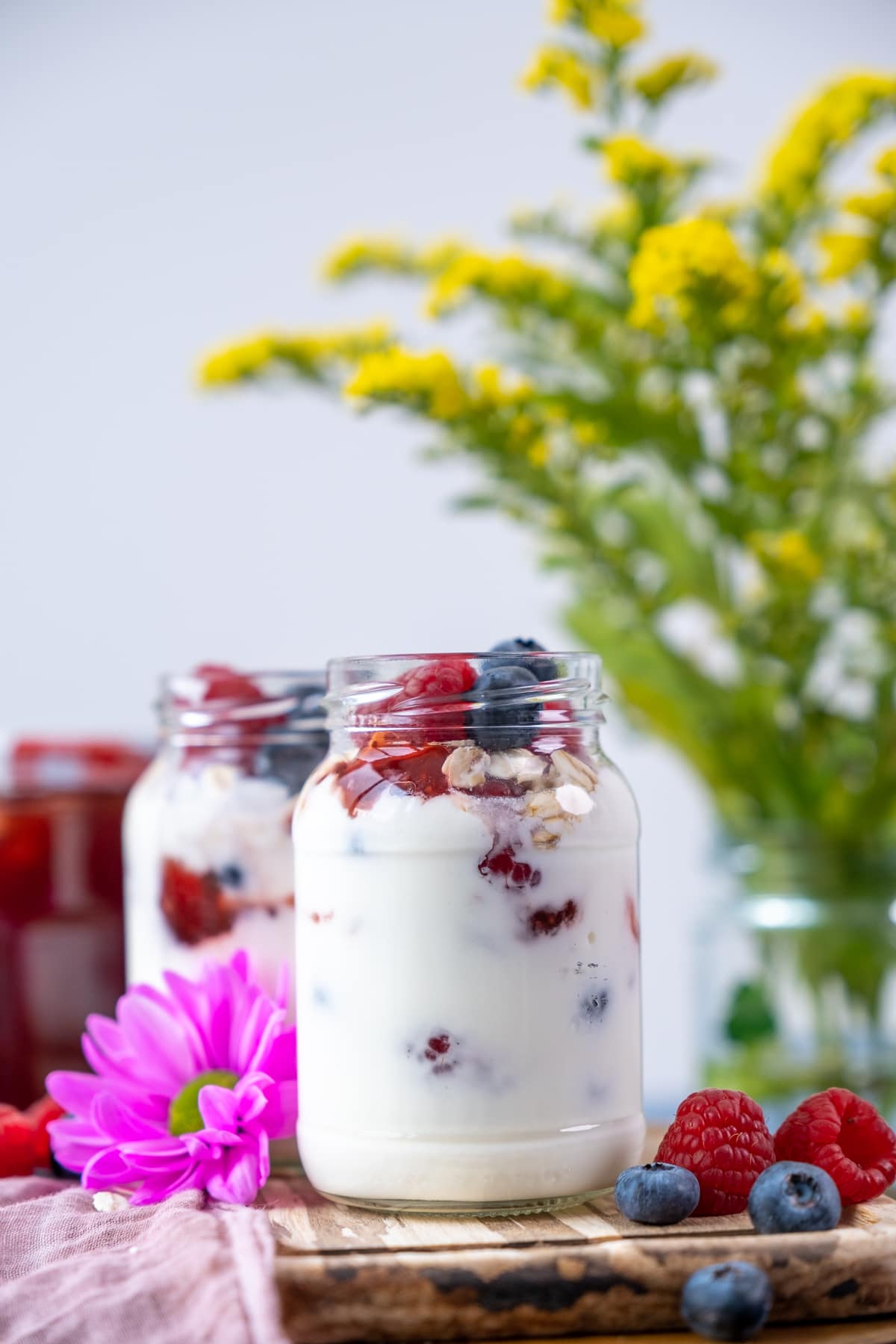 Yogurt parfait with berries in two jars, more berries and a pink flower on the ground and yellow flowers in a vase behind.