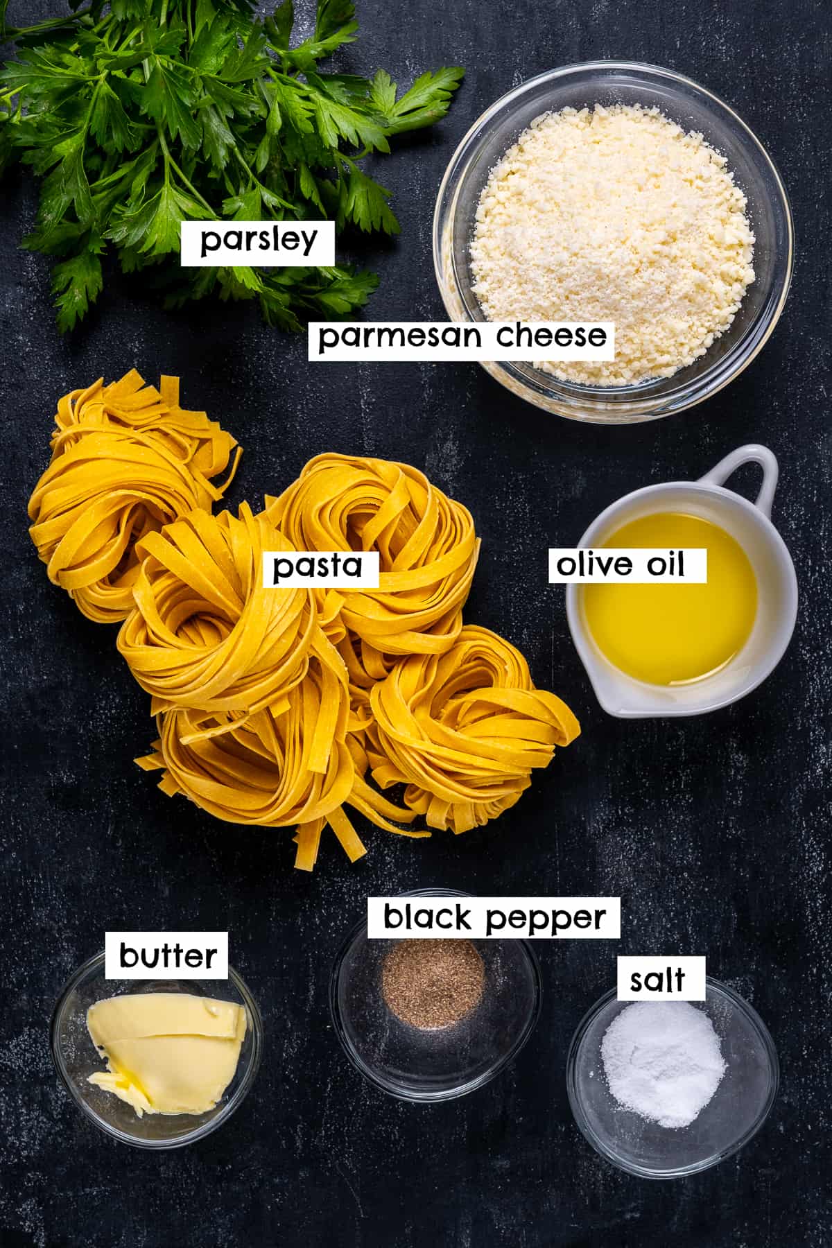 Dry fettuccine, butter, parmesan cheese, oil, parsley, salt and pepper photographed on a black background.