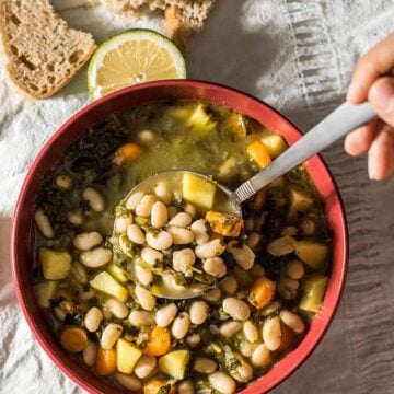 A hand putting white bean soup into a red bowl with a ladle, some crusty bread slices and a wedge of lime on the side.