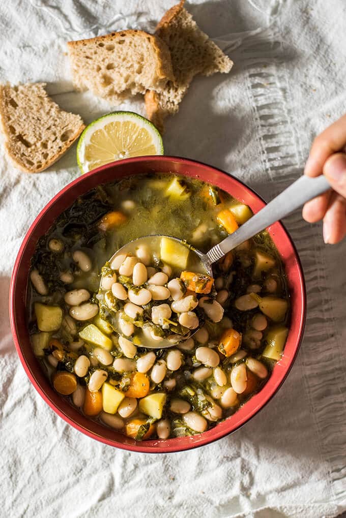 A hand putting white bean soup into a red bowl with a ladle, some crusty bread slices and a wedge of lime on the side.