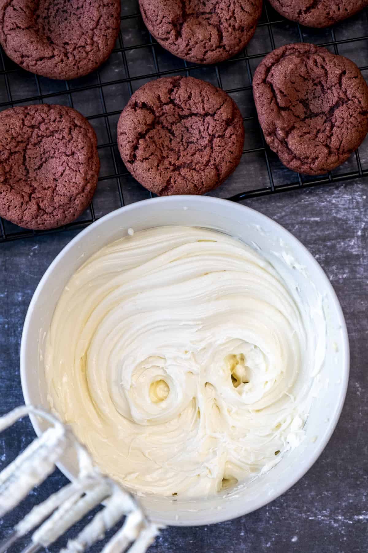 Cream cheese frosting in a bowl, electric mixer and baked red velvet cookies on the side.