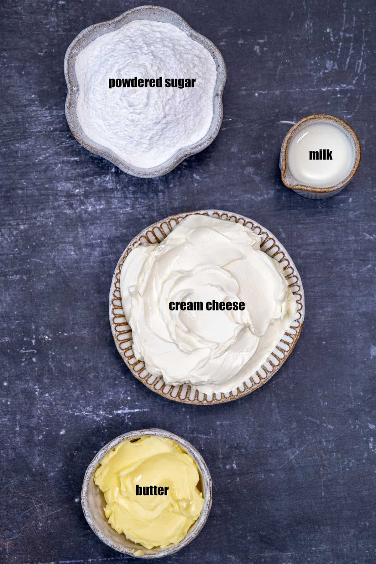 Cream cheese, butter, milk and powdered sugar in separate bowls on a dark background.