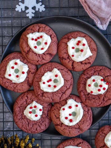 Red velvet cookies with cream cheese frosting decorated with Christmas sprinkles on a black plate.