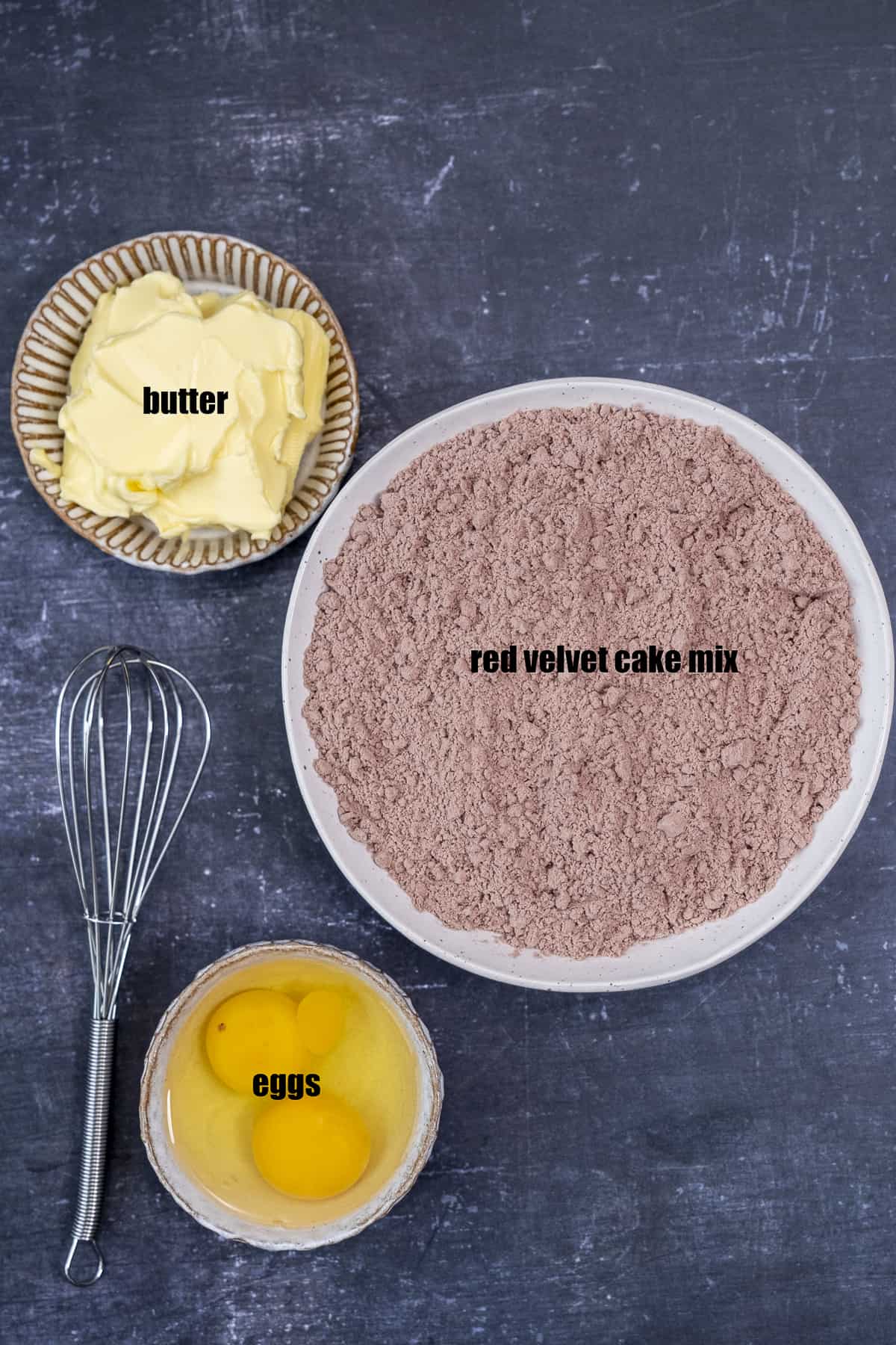 Red velvet cake mix, butter, and 2 eggs without shells in separate bowls and a hand whisk next to these on a dark background.
