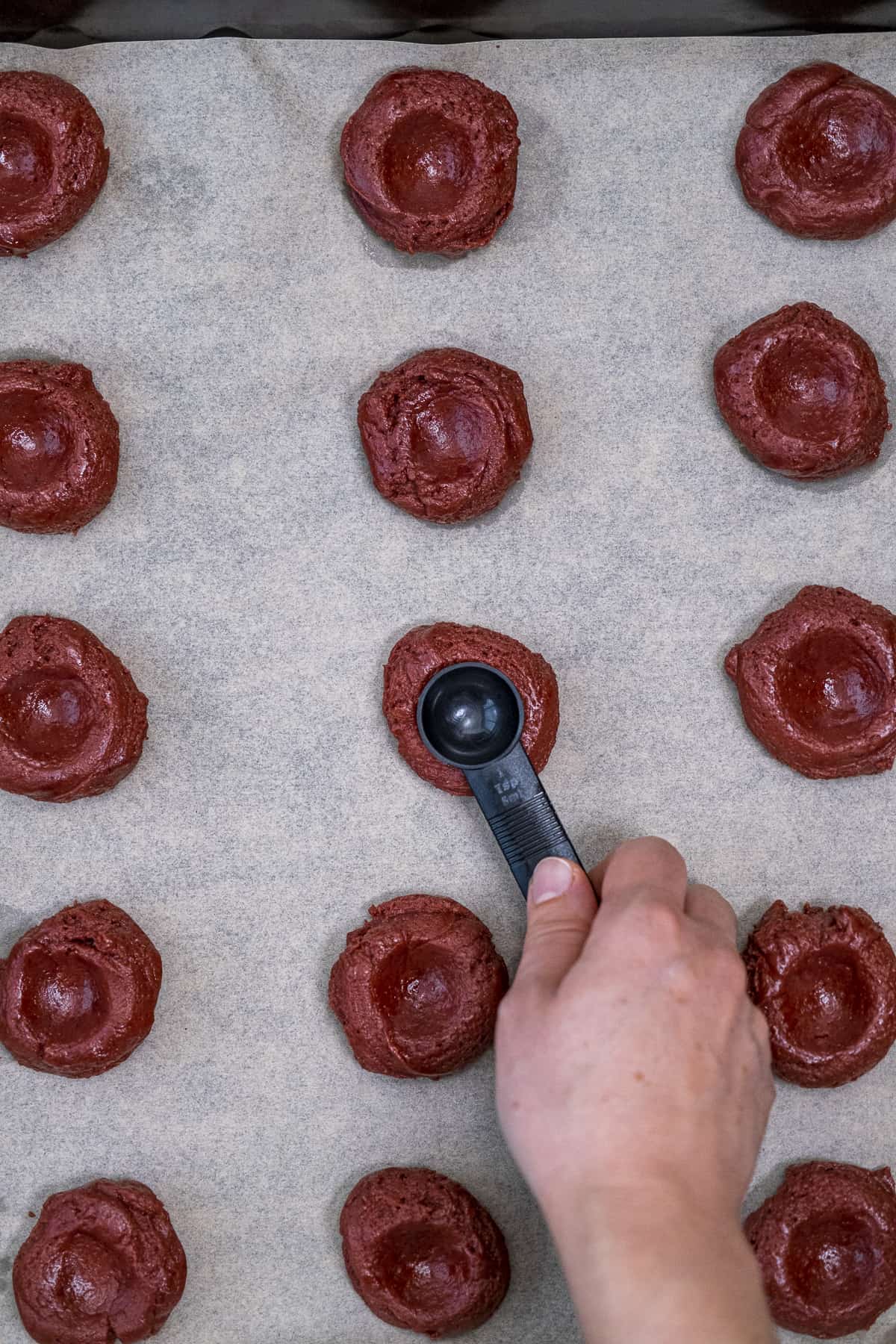 A hand making indentations with a teaspoon on red velvet cookie dough balls on a baking sheet.