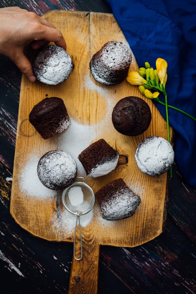 These Buttermilk Chocolate Muffins are perfectly moist, tender and chocolatey. You will feel like you’re eating brownies in each bite. These make a wonderful grab and go breakfast or a quick after school snack. #chocolatemuffins #muffinrecipe #buttermilkmuffins Grabbing one of the Buttermilk Chocolate Muffins dusted with powdered sugar on a wooden board