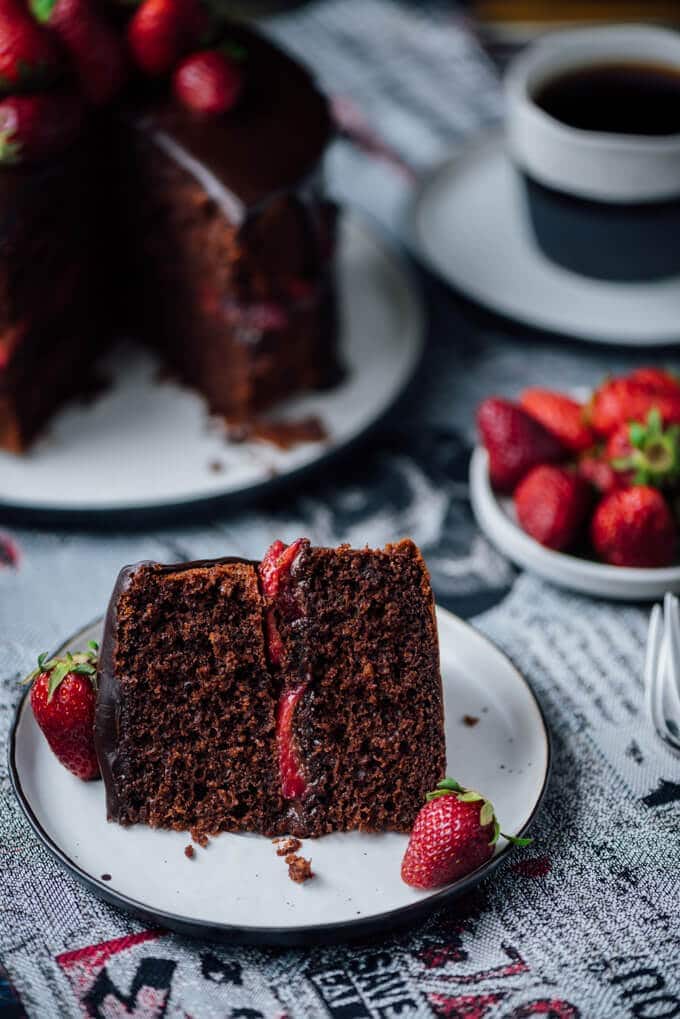 A slice of chocolate cake with strawberry served on a plate. Strawberries and coffee accompany.