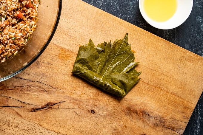 Stuffing and rolling a grape leaf is shown on a wooden board.