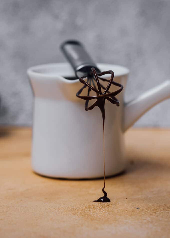 Chocolate ganache dripping from a small hand whisk over a ceramic coffee pot.
