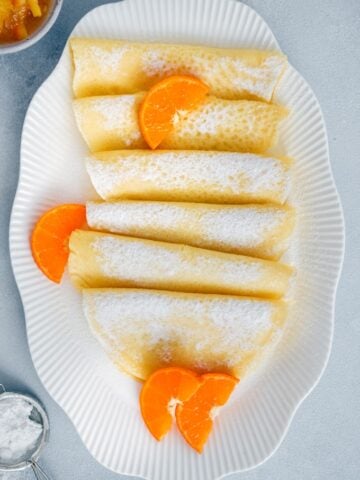 Gluten free crepes served in a white oval plate, garnished with orange slices and powdered sugar.