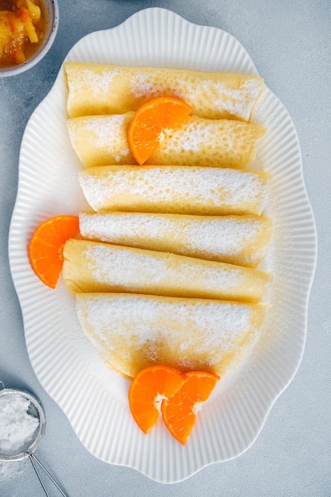 Gluten free crepes served in a white oval plate, garnished with orange slices and powdered sugar.