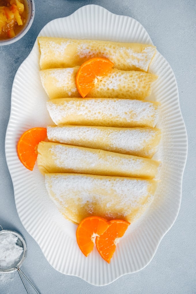 Wonderfully thin gluten free crepes made with rice flour are no different from regular crepes! So easy to make with only 4 simple ingredients. Perfect for several filling options! #glutenfreebreakfast #glutenfreecrepes #riceflourrecipes Gluten free crepes served in a white oval plate, garnished with orange slices and powdered sugar.