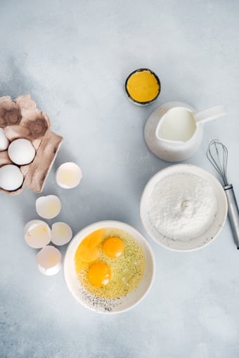 Ingredients for gluten free crepe recipe are photographed in white ceramic bowls on a grey background.