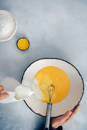 Woman pouring milk into beaten eggs to make a crepe batter in a large ceramic mixing bowl