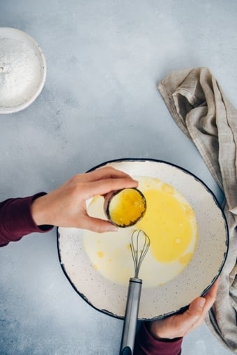 Woman pouring melted butter into the crepe batter in a ceramic mixing bowl.