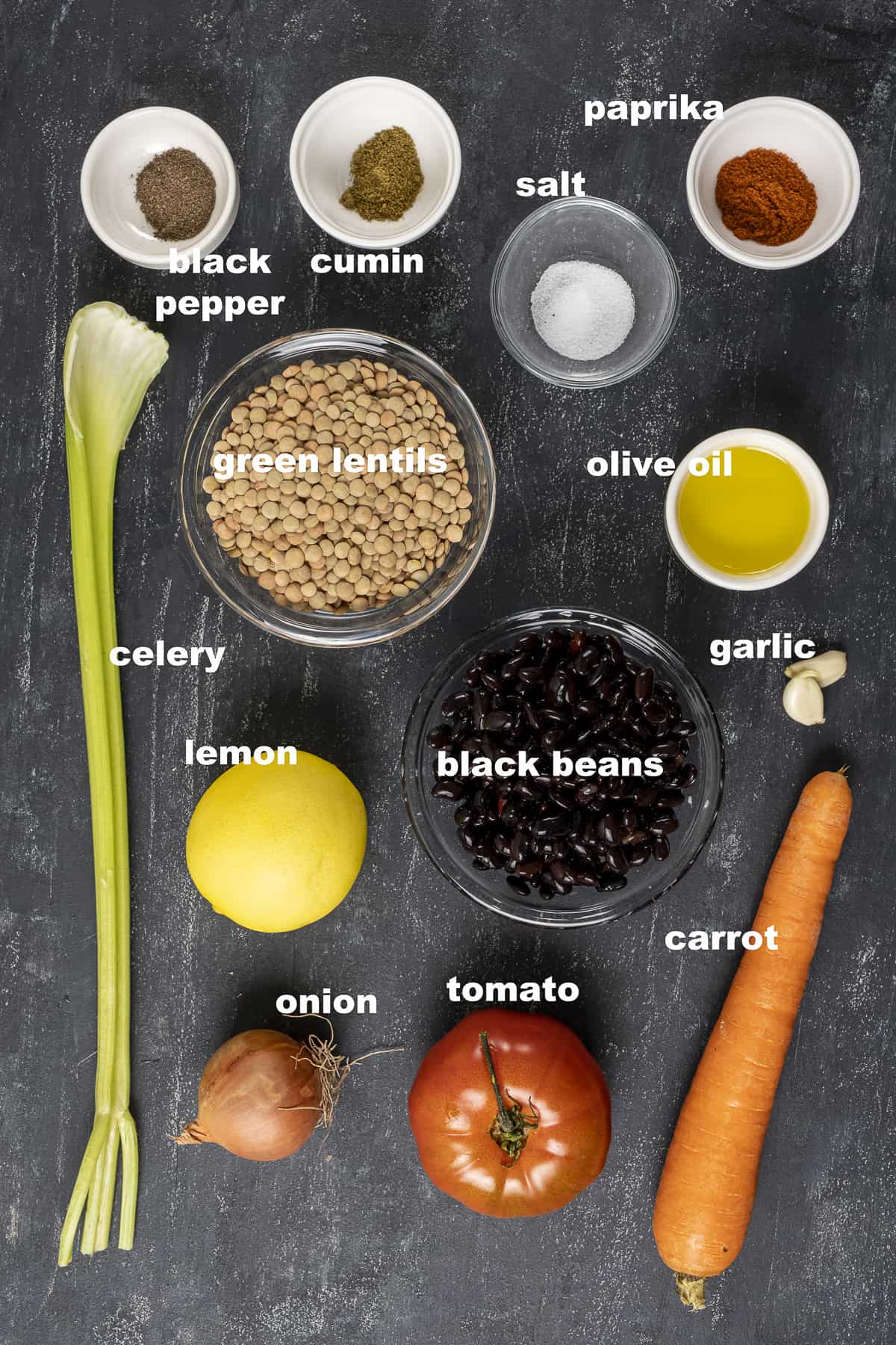 Green lentils, black beans, celery stalk, carrot, onion, tomato, lemon, spices on a dark background.