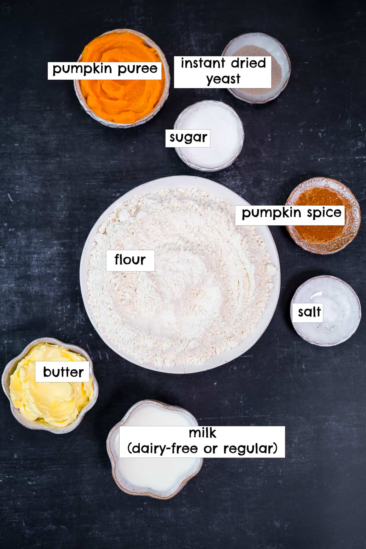 Flour, pumpkin spice, butter, milk, pumpkin puree, sugar and yeast all in separate bowls photographed on a dark background.