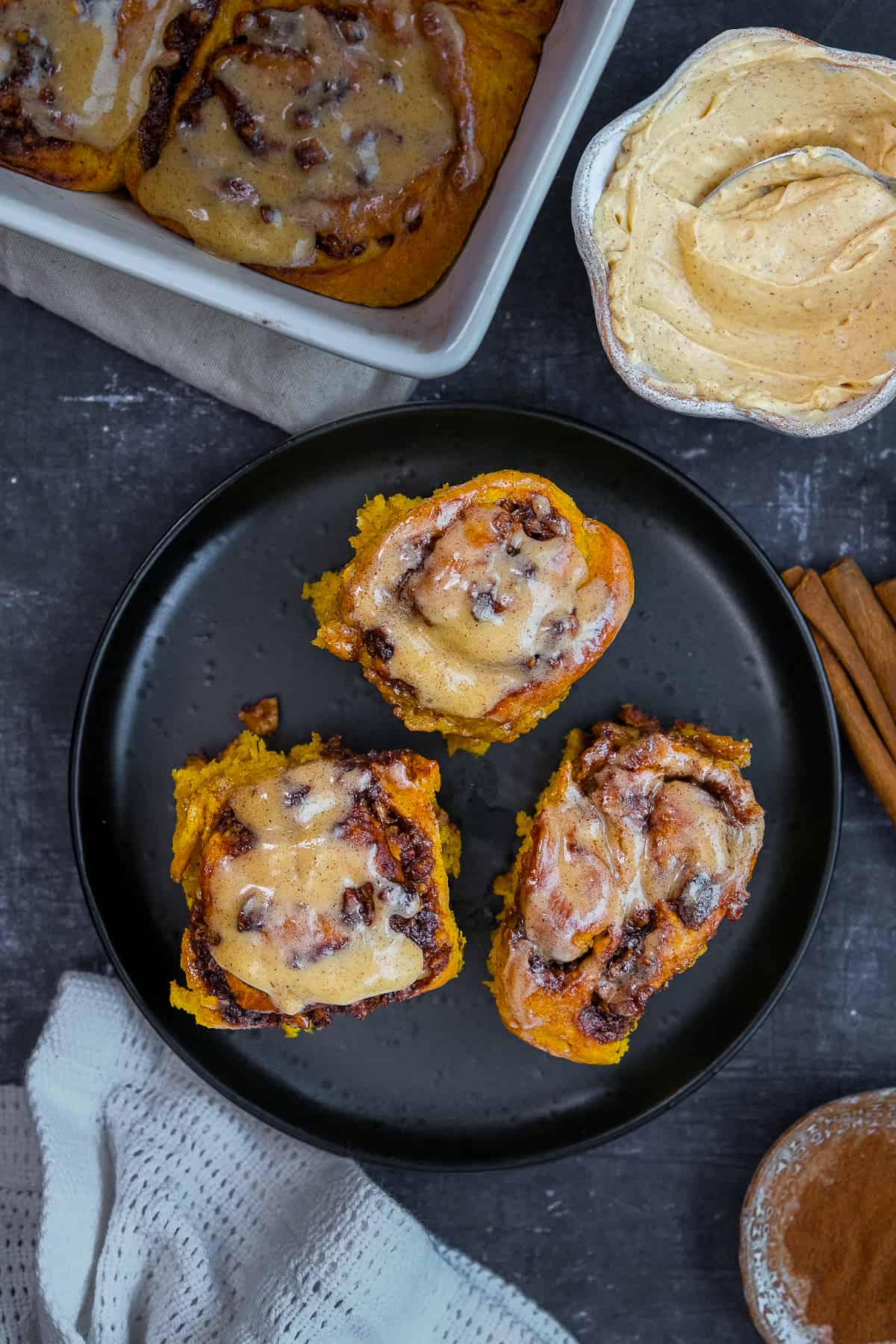 Pumpkin cinnamon rolls with a frosting on their top served on a black plate, cinnamon sticks, baking pan and a bowl of frosting on the side.