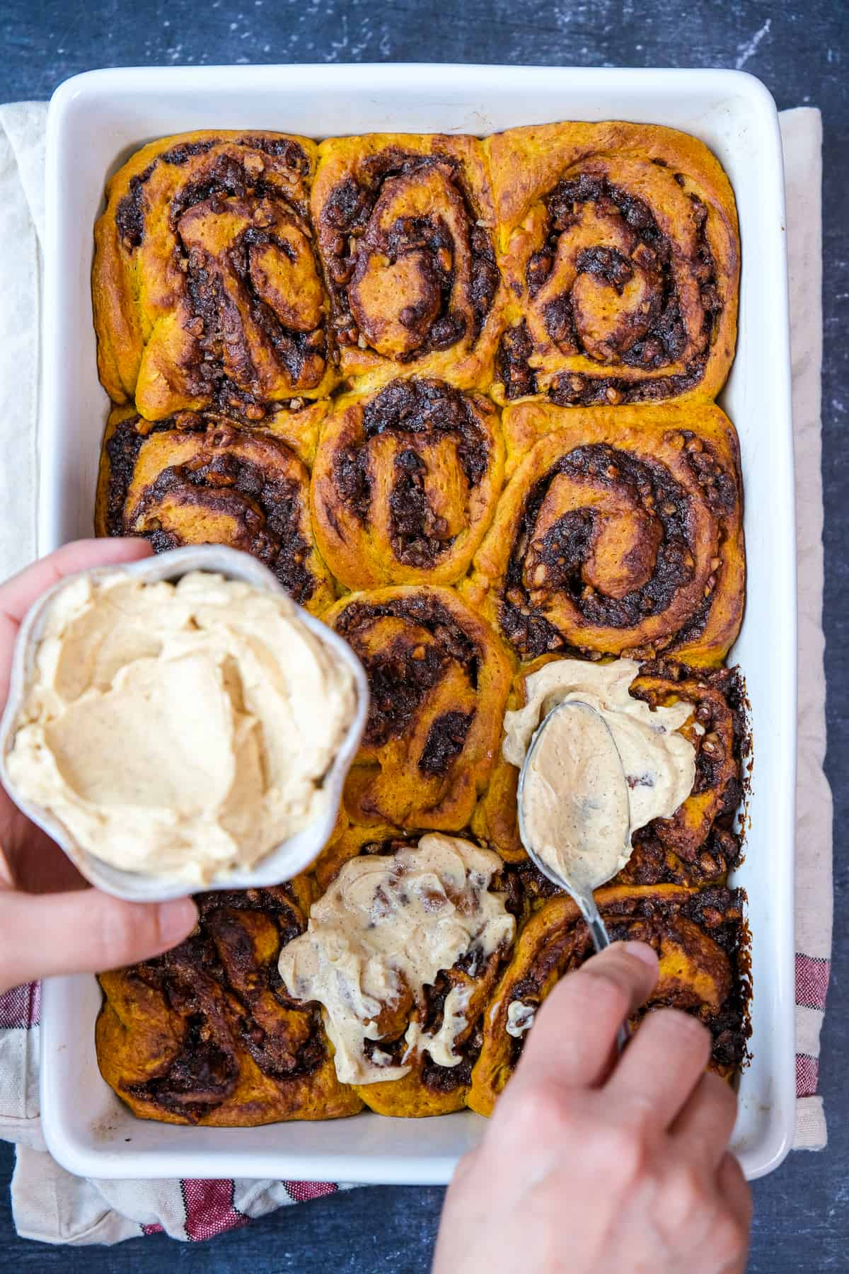 Hands spreading spiced vanilla frosting on baked cinnamon rolls in a baking pan.