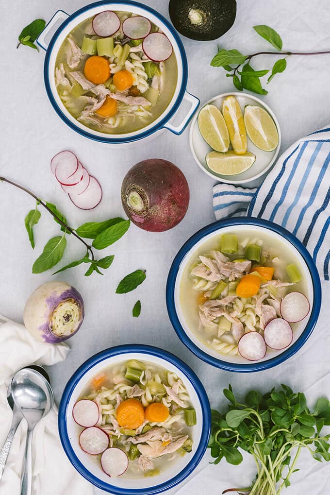 Chicken noodle soup with vegetables served in white bowls and accompanied by radishes and herbs