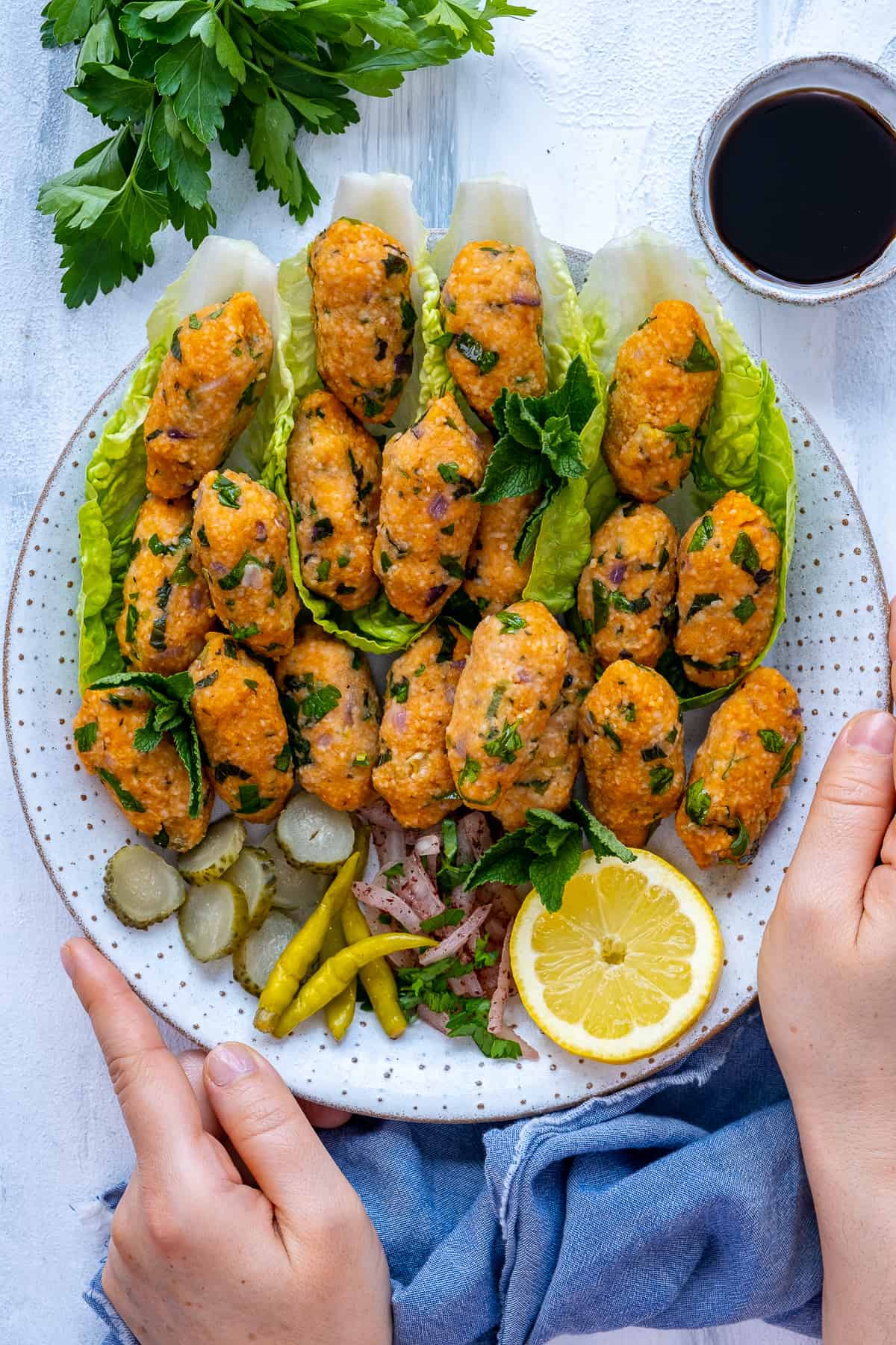 Hands holding a plate full of mercimekli kofte served with lettuce leaves, pickles and onions.