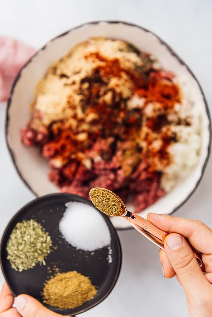 Hands sprinkling spices with a copper teaspoon over kofte ingredients in a ceramic bowl.