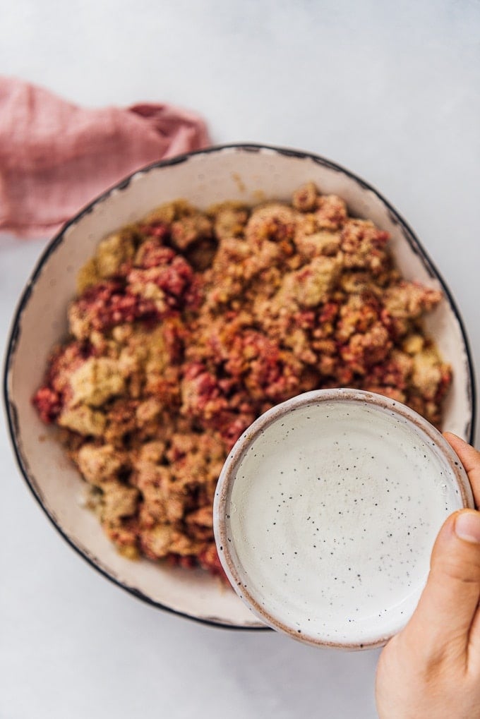 A hand pouring water from a mini ceramic bowl over kofte ingredients.