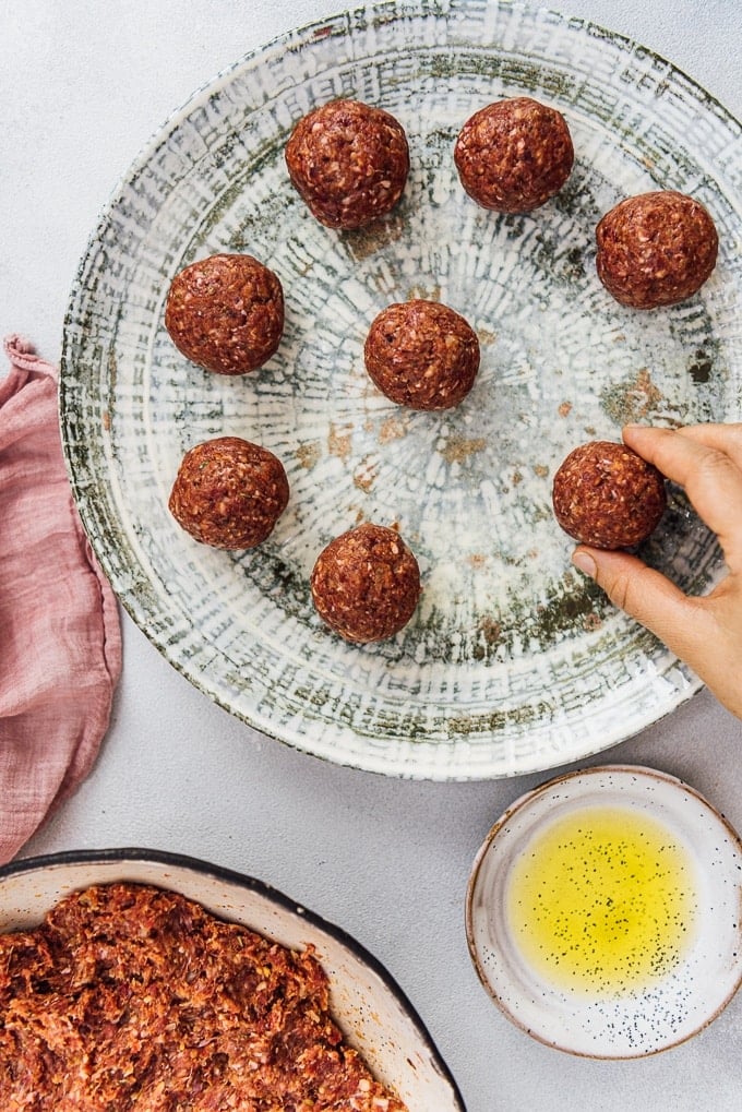 Homemade Turkish Meatballs are spicy, tender and perfectly juicy. It’s not a secret any more to make meatballs at home. #turkishfood #meatballs #meatballsrecipe #middleeasternfood #kofta #kofte A hand placing raw homemade kofte balls on a ceramic plate, olive oil in a mini bowl on the side.