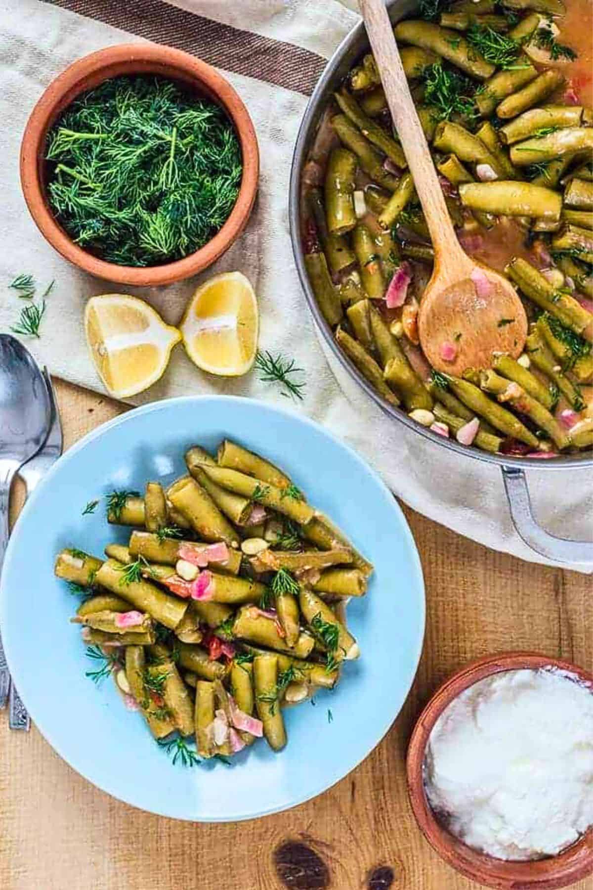 Overhead view of Turkish-style fava beans in pods in a blue bowl, garnished with fresh dill, with a pan of the dish, lemon halves, and yogurt on the side.