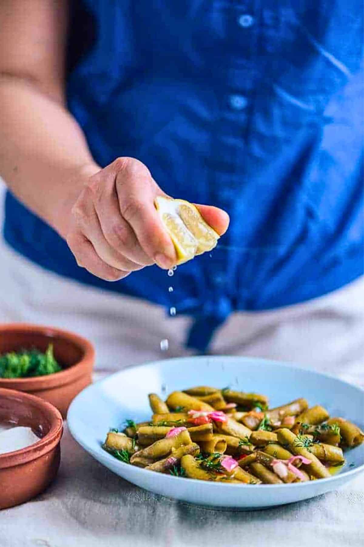 Person squeezing fresh lemon juice over a bowl of Turkish-style fava beans in pods, garnished with dill and onions.