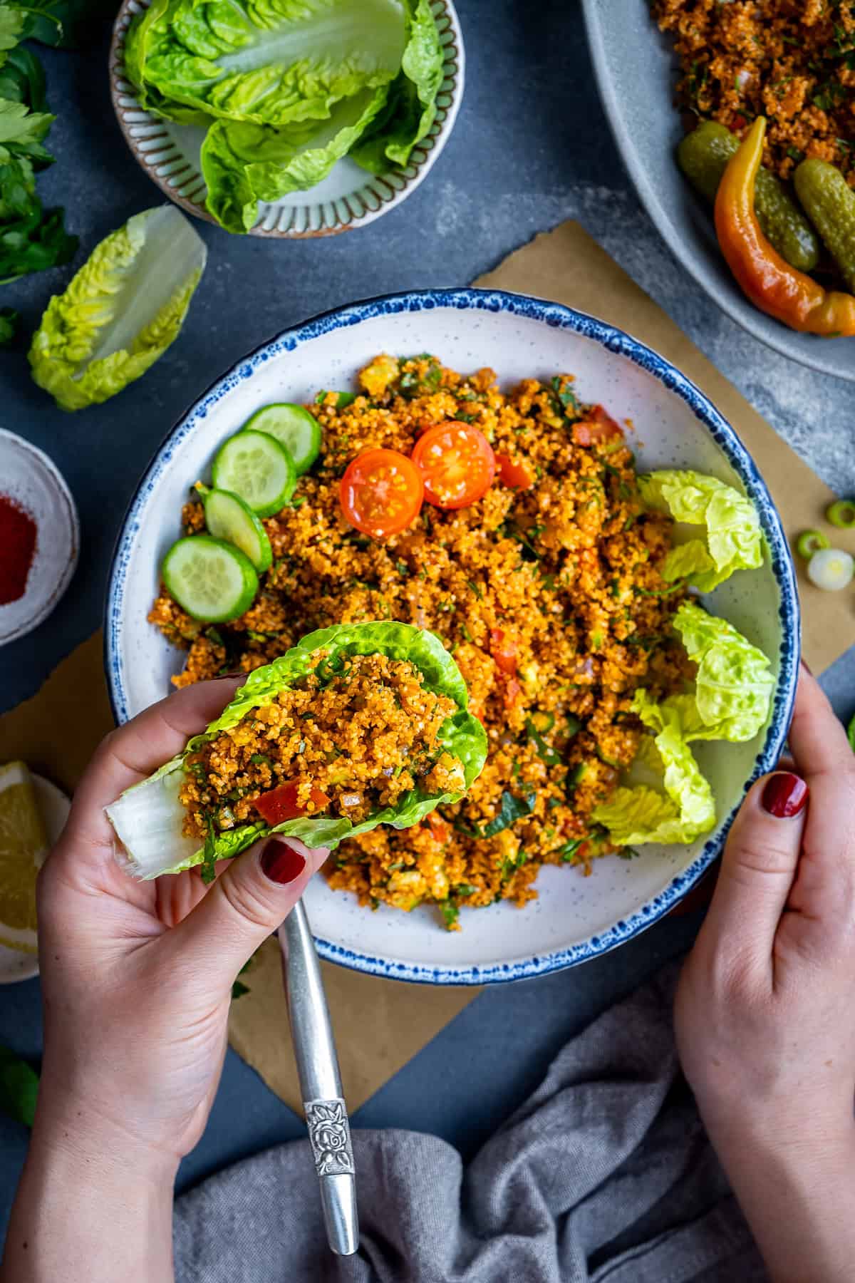 Hand holding a lettuce leaf wrap filled with Turkish kisir, served with a bowl of bulgur salad, cucumbers, tomatoes, pickles, and lettuce leaves.