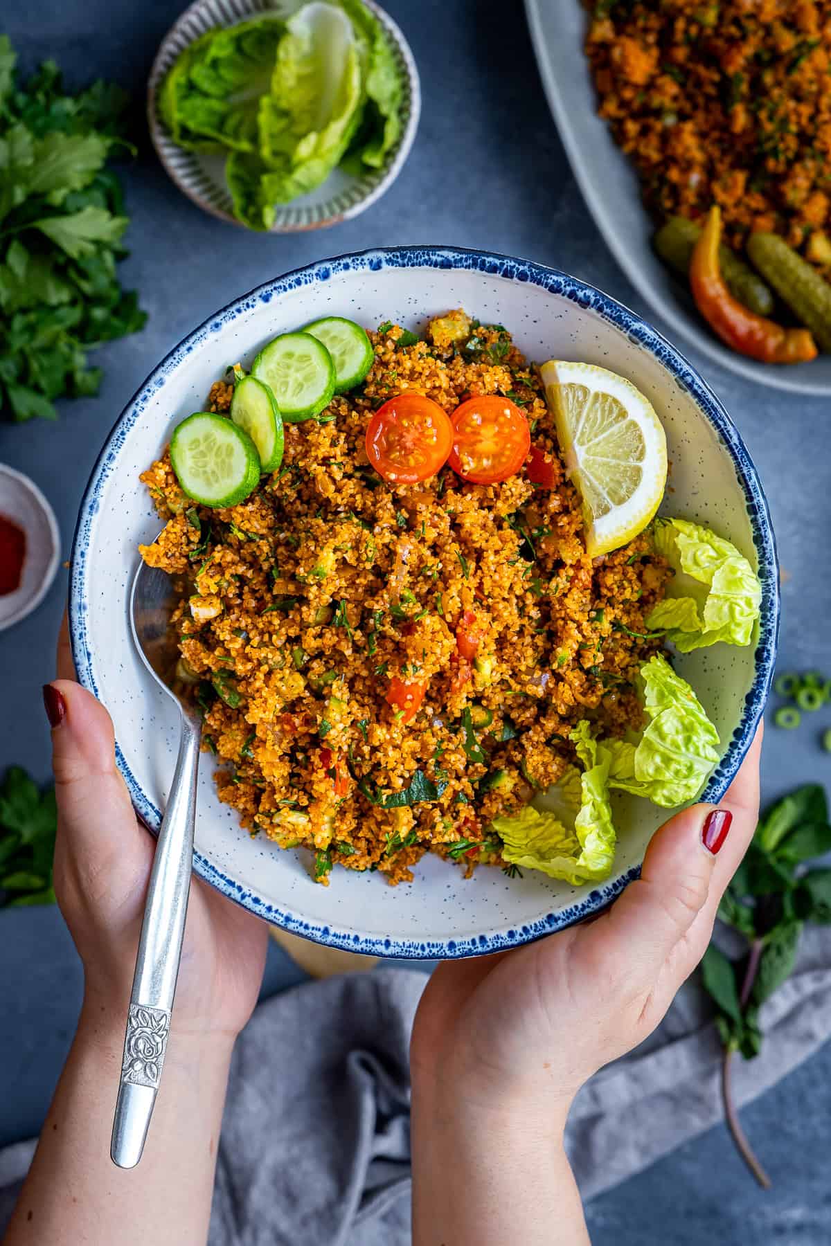 Hands holding a bowl of Turkish kisir, a bulgur salad with herbs, tomatoes, cucumbers, lemon wedges, and lettuce leaves on the side.