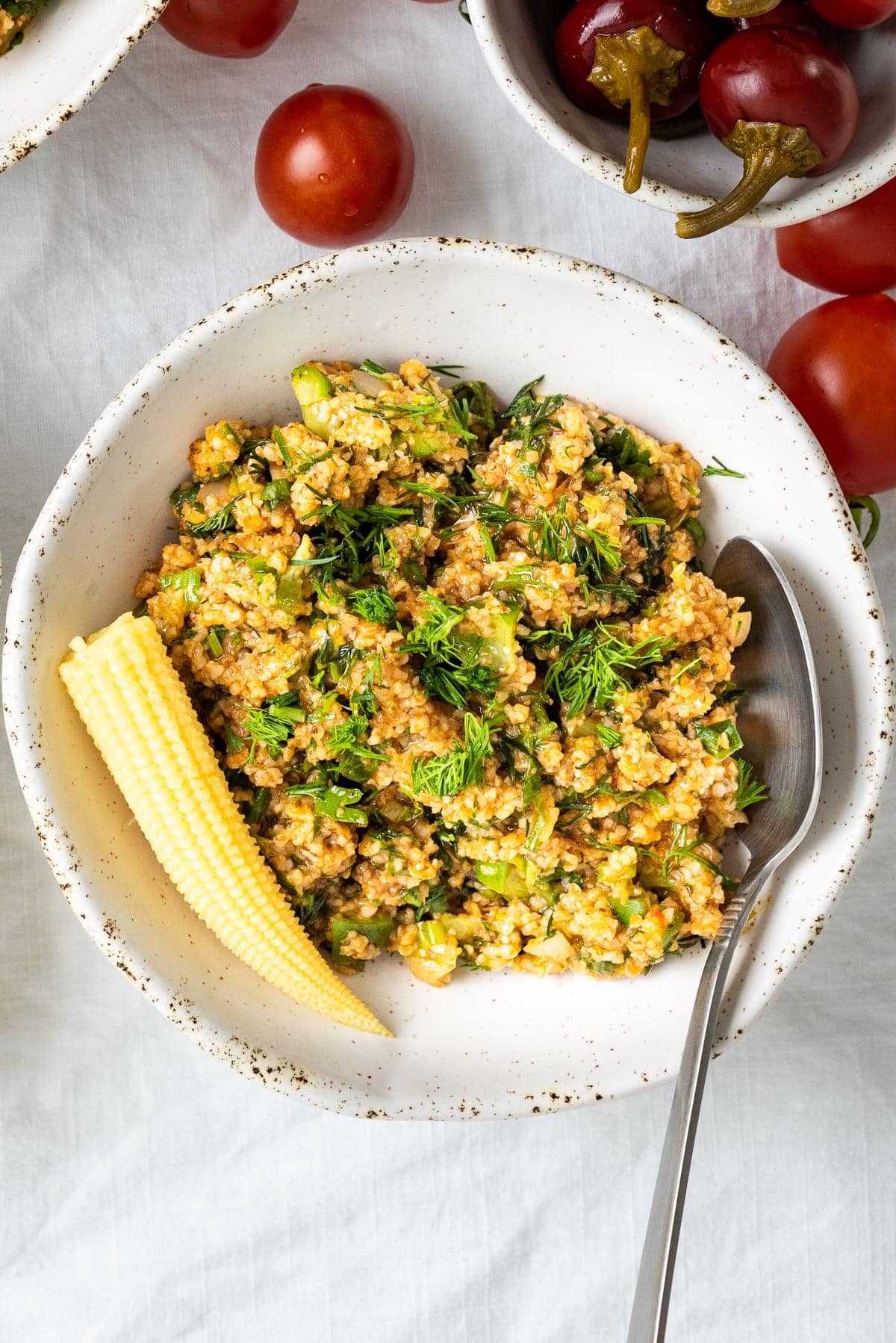 Turkish bulgur salad kısır in a white ceramic bowl, a spoon inside it and a pickled baby corn on the left top.