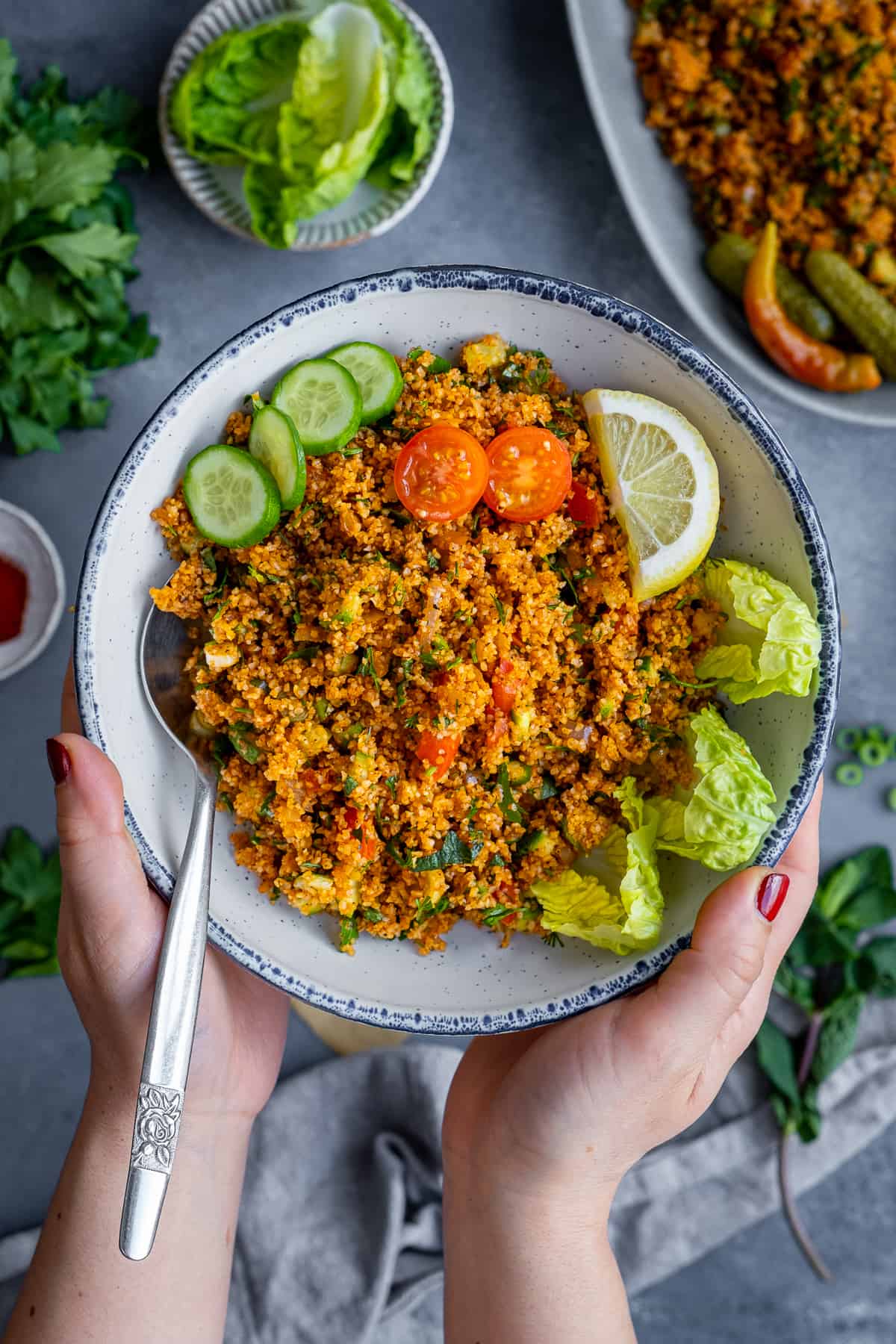 Hands holding a bowl of bulgur salad garnished with lettuce leaves, a lemon wedge, sliced tomatoes and cucumbers.