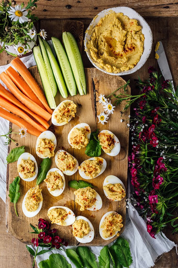 Healthy Deviled Eggs with hummus are on a snack board accompanied by cucumber and carrot sticks.