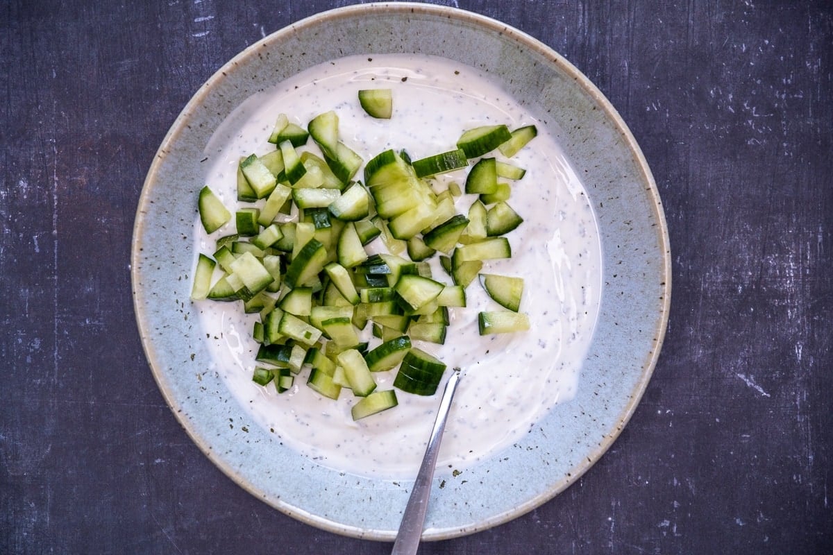 Chopped cucumber on the yogurt, salt and dried mint mixture in a large bowl and a spoon inside it.