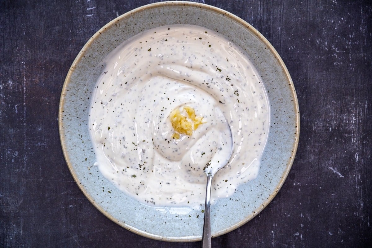 Mashed garlic in the yogurt, salt, dried mint mixture in a blueish bowl and a spoon inside it.