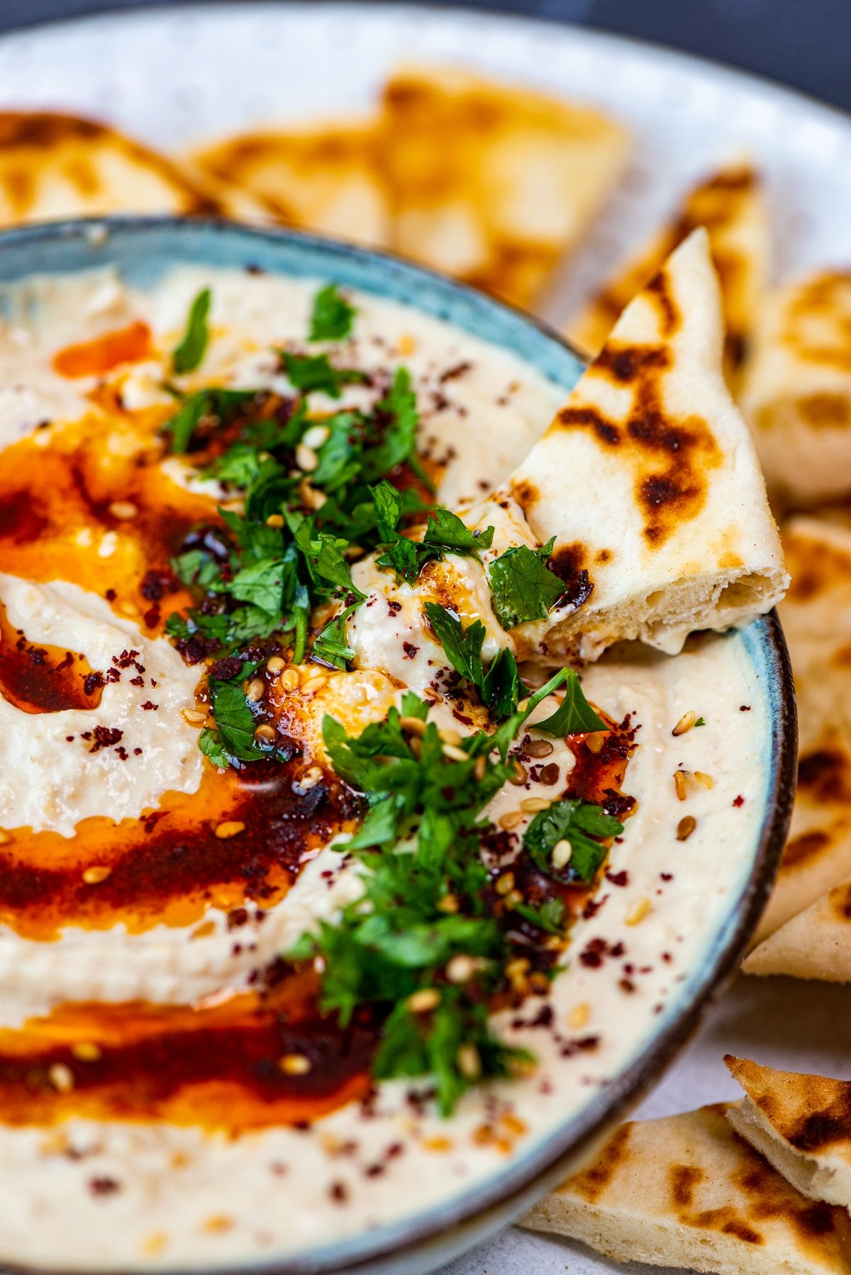 Homemade hummus in a bowl topped with paprika oil, parsley and sumac. A piece of bread in it.