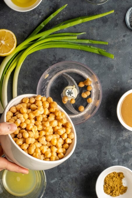 A hand putting cooked chickpeas from a white bowl into a food processor. Green onions, lemon, tahini and spices are on the side.