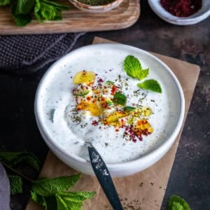 A bowl of Turkish cacık topped with olive oil, diced cucumber, dried mint, pul biber, sumac and fresh mint.