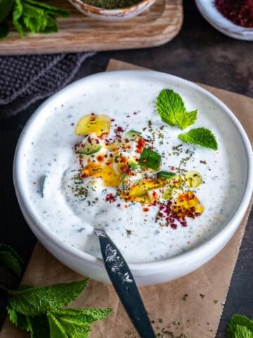A bowl of Turkish cacık topped with olive oil, diced cucumber, dried mint, pul biber, sumac and fresh mint.