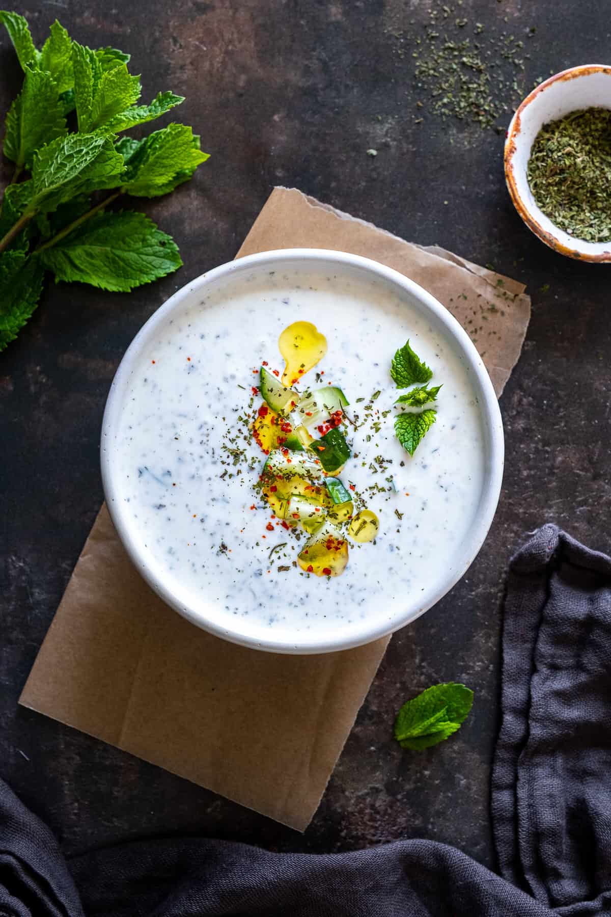 Overhead view of a bowl of cacık topped with olive oil, diced cucumber, dried mint, pul biber and fresh mint leaves.
