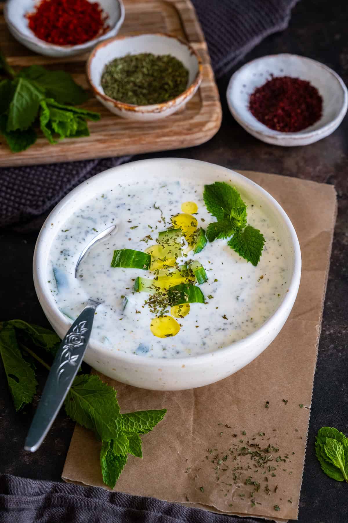 A bowl of cacık topped with olive oil, cucumber slices and fresh mint, with a spoon and bowls of dried mint, pul biber and sumac in the background.
