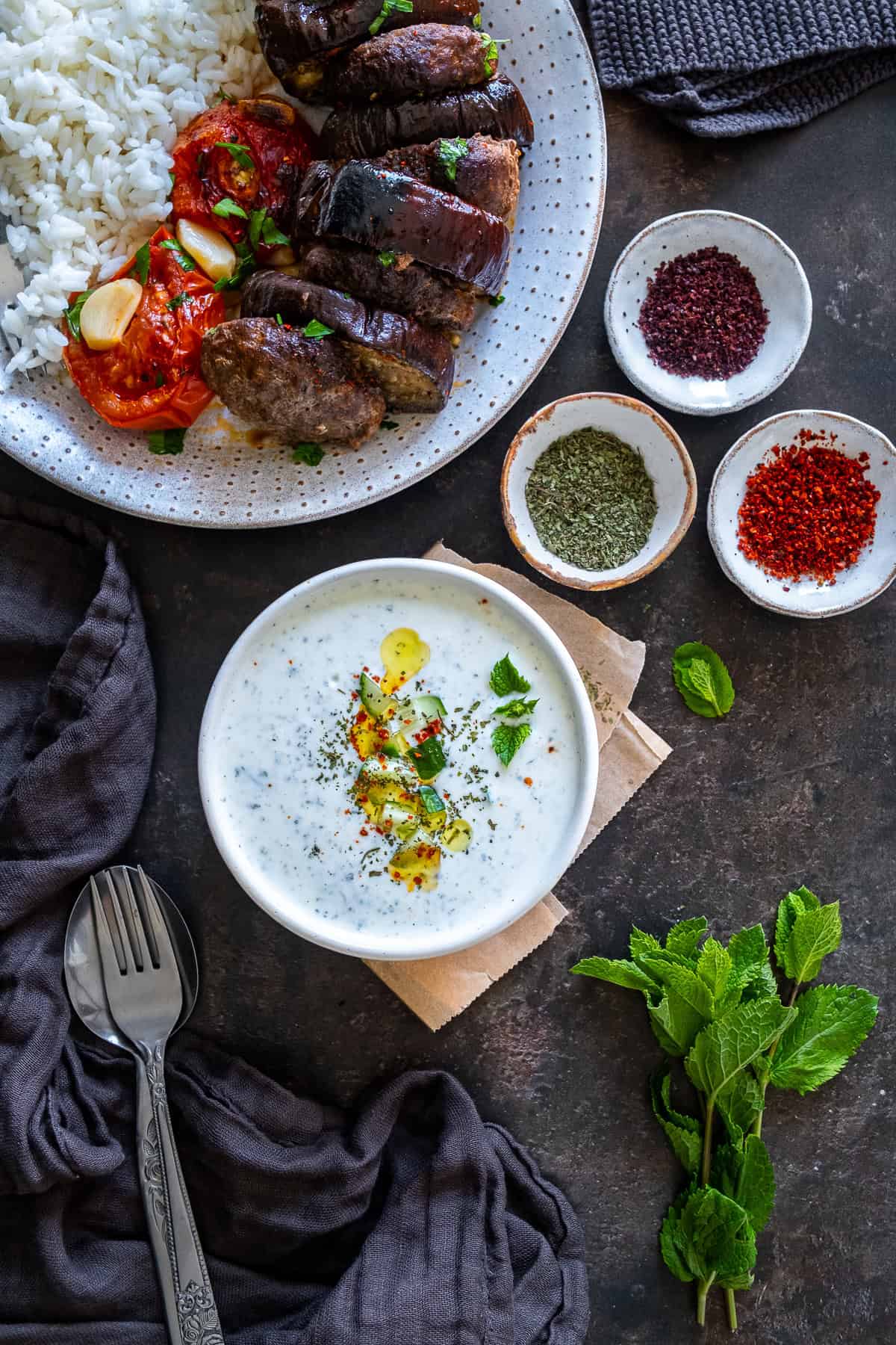 A bowl of cacık served with Turkish eggplant kebab and rice pilaf, with bowls of dried mint, pul biber and sumac on the side.