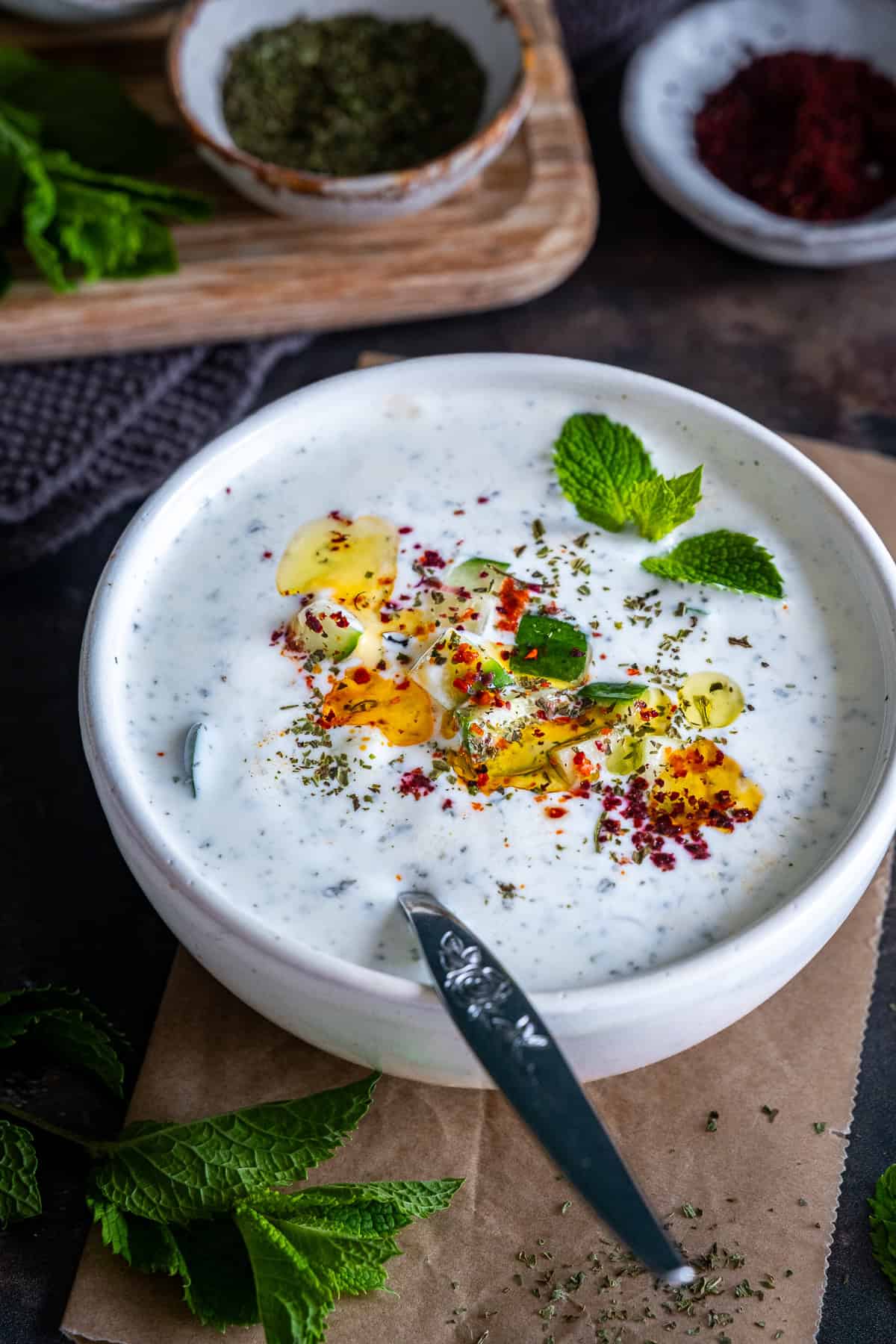 Close-up of a bowl of cacık topped with olive oil, diced cucumber, dried mint, pul biber, sumac and fresh mint leaves.