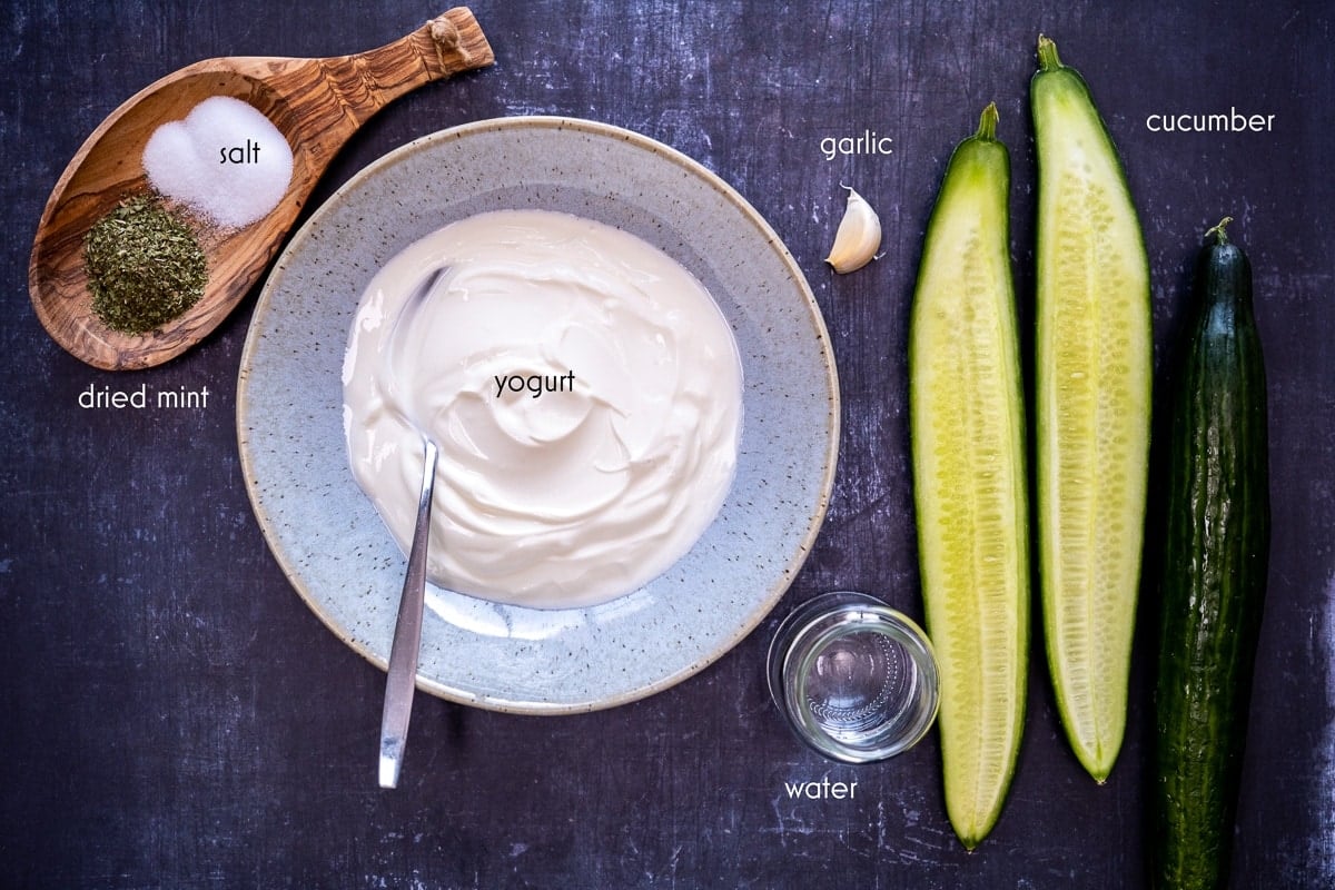 Yogurt in a large bowl, halved cucumber, a whole cucumber, garlic, water in a small bottle, salt and dried mint in a wooden bowl on a dark background.