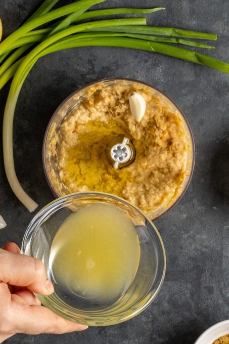 A hand pouring lemon juice from a small glass bowl over mashed chickpeas in a food processor.