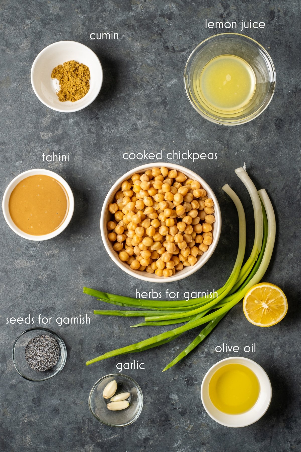 Cooked chickpeas, lemon juice, olive oil, salt and cumin, tahini, garlic cloves in separate bowls and green onions on the ground from top view.