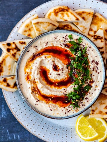 Authentic hummus served in a bowl topped with paprika oil, sumac and parsley. Pide bread chunks and lemon wedges on the side.