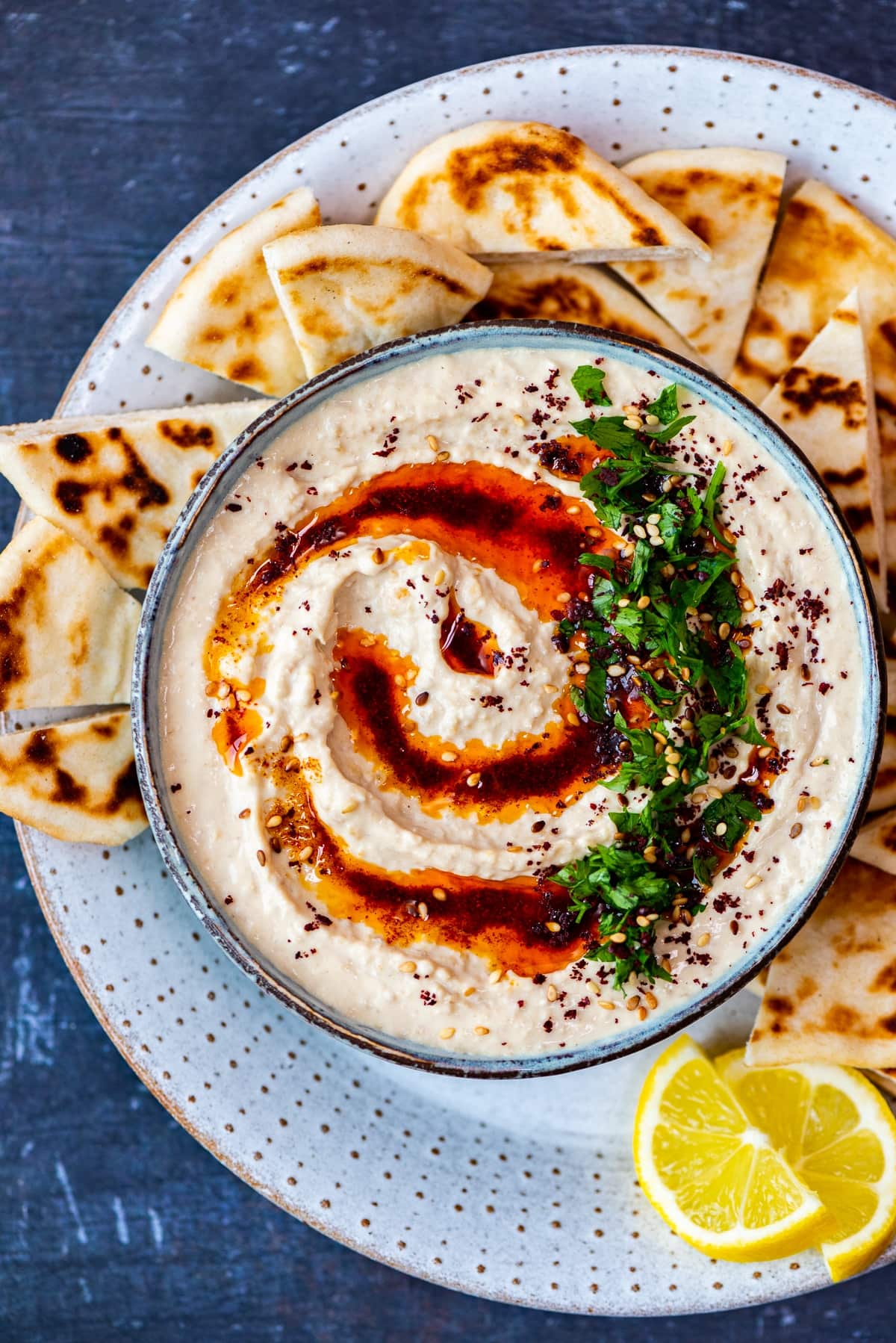 Authentic hummus served in a bowl topped with paprika oil, sumac and parsley. Pide bread chunks and lemon wedges on the side.